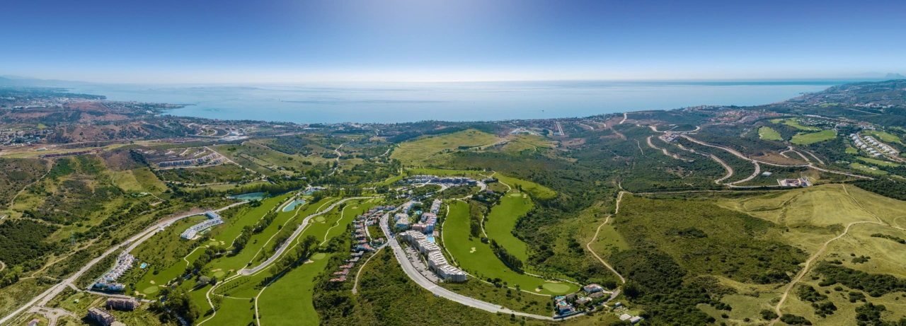 En omfattende luftfoto af Sunny Golf-udviklingen, perfekt integreret med de frodige grønne fairways på Estepona Golf og det funklende Middelhav i baggrunden.